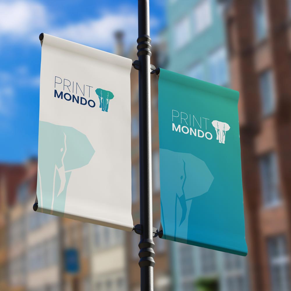 PVC Banner mit Hohlsaum für Außenwerbung, an einer Metallstange an der Straße. Hier mit Beispieldruck in weiss und türkis mit dem Print Mondo Logo und dem Elefanten.