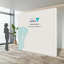 Messewand Basic, leichtes Textilbanner für ihren Firmenauftritt in einem Büro neben einer Person. Hier mit einem Beispieldruck in weiss und türkis mit dem Print Mondo Elefanten Logo in blau.