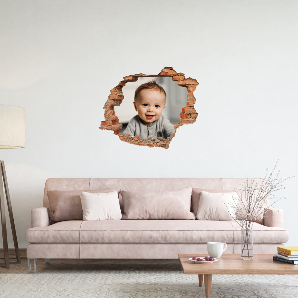 Ein Wandaufkleber, das durch einen Mauerbruch betrachtet wird, zeigt ein Lächeln des Babys in einem grauen Outfit. Das Bild ist an einer Wand in einem Wohnzimmer über einem rosa Sofa mit Kissen und einem Tisch mit einer Tasse und Keksen platziert.