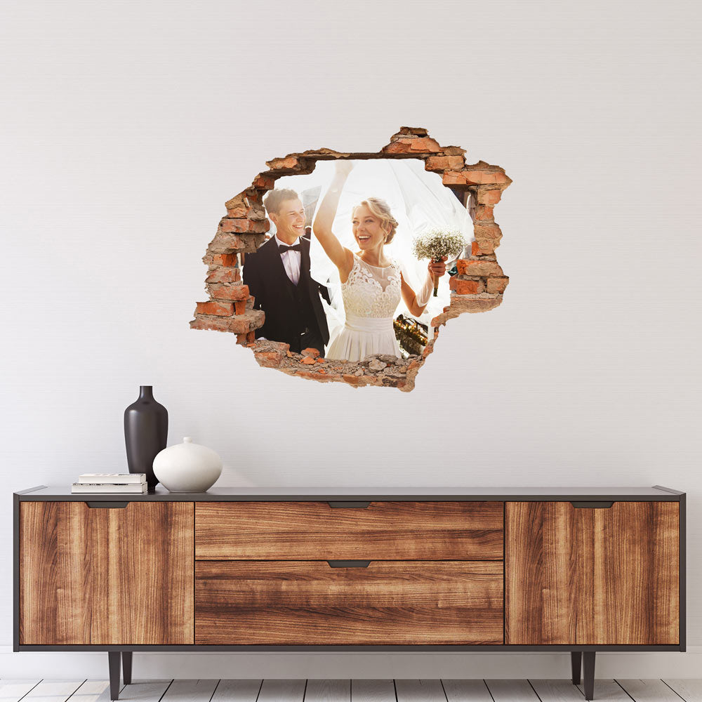Ein Hochzeitsfoto von einem glücklichen Brautpaar, das durch einen Mauerbruch auf einer Wand gesehen wird. Die Braut trägt ein weißes Hochzeitskleid, während der Bräutigam einen schwarzen Anzug trägt. Sie halten ihre Hände und die Braut hält ihren Brautstrauß. Das Bild ist an einer Wand über einer Holzkommode platziert.