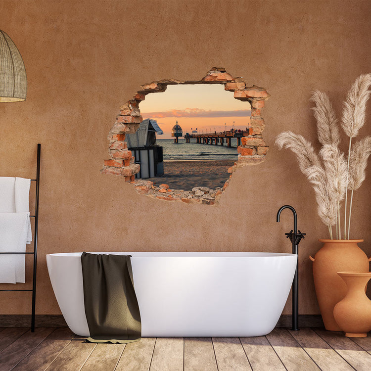 Ein wunderschöner Blick auf einen Strandpier mit Sonnenuntergang, gesehen durch einen Mauerbruch-Aufkleber in einer Wand. Der Pier erstreckt sich in den Horizont, umgeben von einem goldenen Sonnenuntergang. Im Vordergrund ist eine Badewanne in einem modernen Badezimmer mit Pflanzen und einem Handtuchhalter zu sehen.