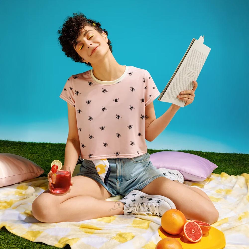 Junge Frau entspannt auf einer Picknickdecke mit einem Getränk und einem Buch. Sie trägt ein rosa Crop-T-Shirt mit schwarzem Bienenmuster, kombiniert mit Jeansshorts und passenden Sneakers. Perfekter sommerlicher Freizeitlook.