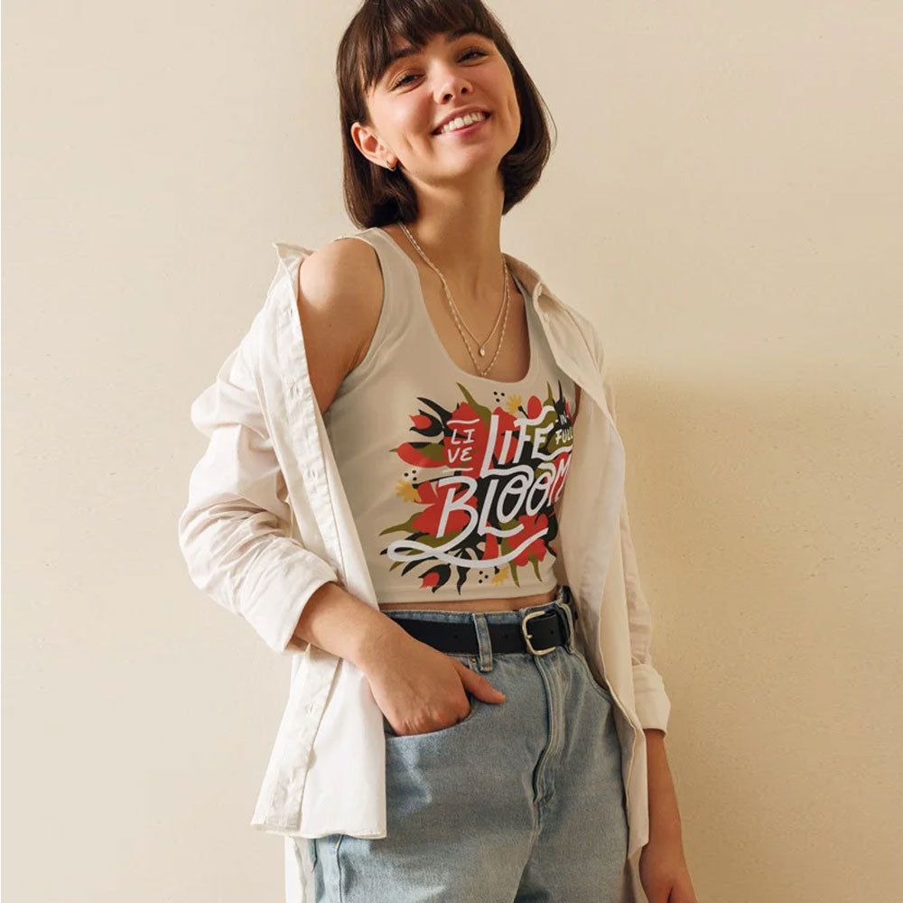 Junge Frau mit kurzen braunen Haaren trägt ein beiges Crop-Top mit farbenfrohem 'Live Life Bloom'-Print, kombiniert mit einer offenen weißen Bluse und einer hoch geschnittenen Jeans. Perfekt für lässige Streetwear und sommerliche Outfits