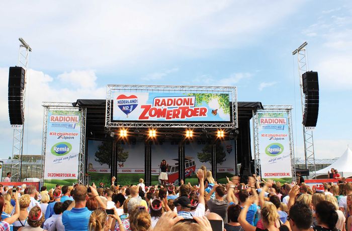 Open-Air-Bühne beim RadioNL Zomertoer Musikfestival in den Niederlanden. Eine große Menschenmenge feiert vor der Bühne, während ein Künstler auftritt. Die Bühne ist mit Bannern und dem Schriftzug 'RADIONL ZOMERTOER' dekoriert, flankiert von Sponsorenlogos. Das Event findet unter freiem Himmel bei sonnigem Wetter statt.