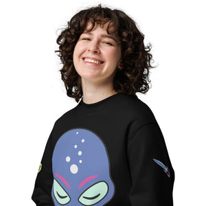 Eine Frau trägt ein schwarzes Sweatshirt mit einem auffälligen Alien-Print auf der Vorderseite. Das Design zeigt einen blauen Alien-Kopf mit geschlossenen Augen, rosa Akzenten und kleinen Blasen, die einen futuristischen Look erzeugen. Zusätzlich befindet sich auf dem Ärmel eine kleine bunte Rakete als weiteres Detail des individuellen Drucks. Das Sweatshirt hat eine lockere Passform und besteht aus weichem Material, ideal für ein bequemes und modernes Outfit.