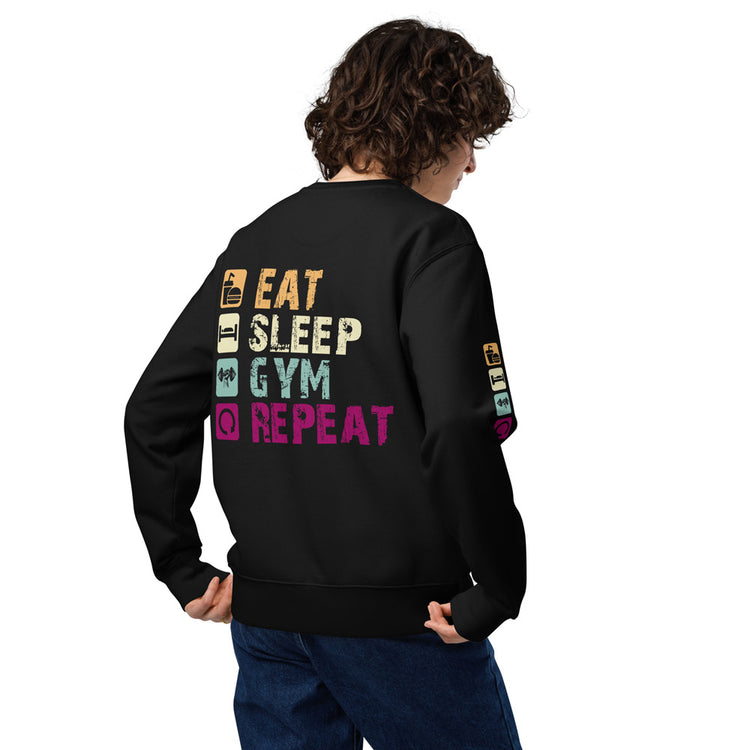 Eine Frau trägt ein schwarzes Sweatshirt mit einem auffälligen Fitness-Motiv auf dem Rücken. Der bunte Print zeigt die Wörter "Eat Sleep Gym Repeat" in einem coolen Vintage-Stil mit dazugehörigen Symbolen für Essen, Schlafen, Fitnessstudio und Wiederholung. Auch auf den Ärmeln befinden sich kleine Icons, die das Design abrunden. Das Sweatshirt hat eine bequeme Passform, elastische Bündchen und besteht aus weichem Stoff – perfekt für Sportliebhaber oder als lässiges Freizeit-Outfit.