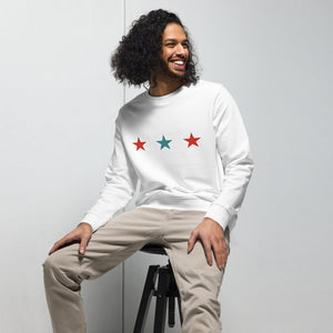 Junger Mann mit lockigem Haar trägt ein weißes Sweatshirt mit drei Sternen in Rot und Blau auf der Brust. Er sitzt entspannt auf einem schwarzen Hocker, kombiniert mit beigefarbener Hose. Perfekter minimalistischer und moderner Streetwear-Look.