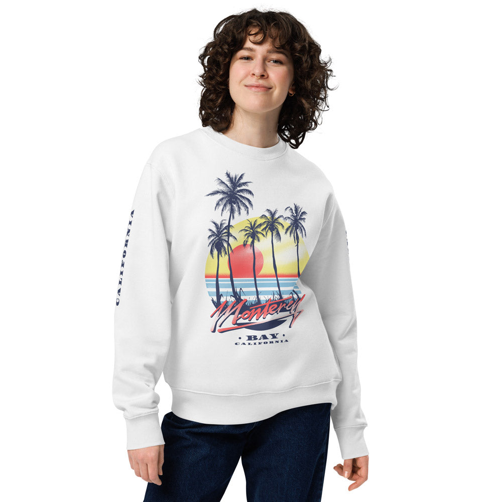 Eine Frau trägt ein weißes Sweatshirt mit einem großen, farbenfrohen Monterey-Bay-Print auf der Vorderseite. Das Design zeigt eine tropische Strandszene mit Palmen, Sonnenuntergang und dem Schriftzug "Monterey Bay California" in einem Retro-Stil. Die Ärmel sind mit zusätzlichem Text-Print verziert. Das Sweatshirt hat eine bequeme Passform mit gerippten Bündchen und ist aus weichem Stoff gefertigt – ideal für lässige und stilvolle Outfits.